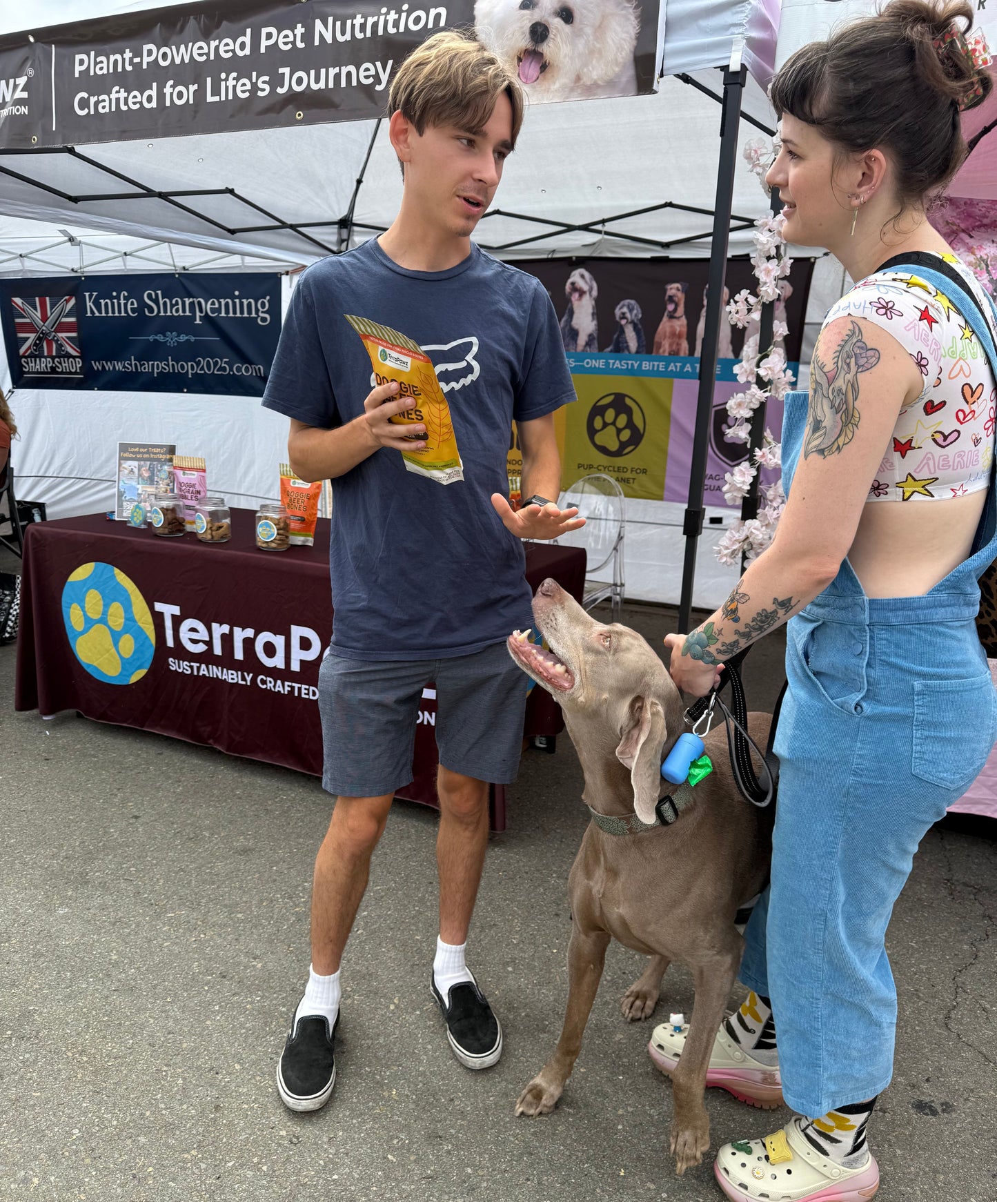 Two people with a dog at an outdoor event with TerraPawz treats displayed.