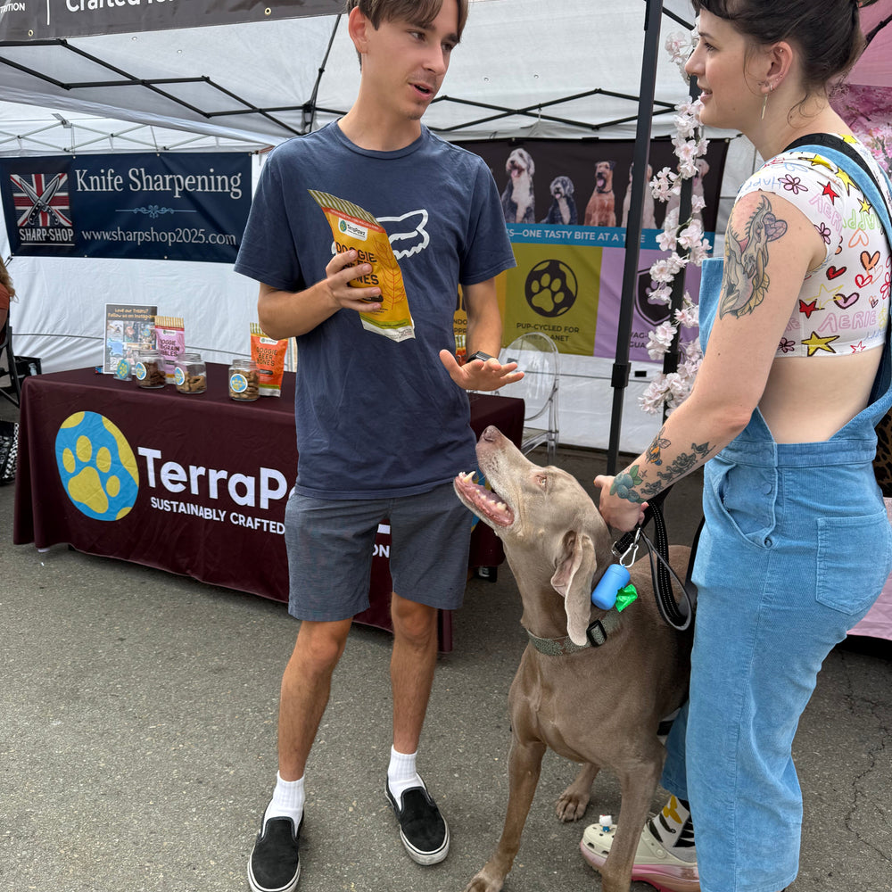Two people with a dog at an outdoor event with TerraPawz treats displayed.